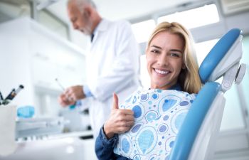 Dentist and a satisfied young woman with a perfect smile sitting in a dental chair and showing her thumb up.