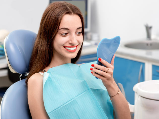 A satisfied young woman in a dental chair looking at her new perfect smile in a mirror.