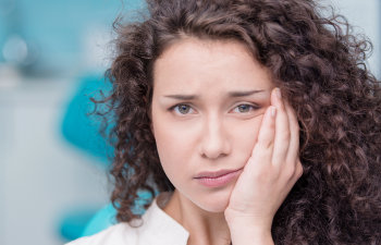 Woman with curly hair looking worried, holding her cheek, with a blurred blue background.